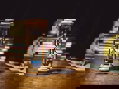 Wooden hourglass with blue sand on a desk, with books and office items blurred in the background.