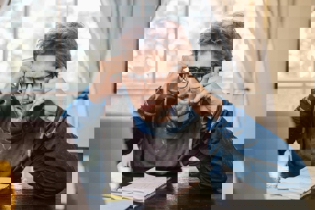 Frustrated man wearing glasses talks on the phone while looking at a laptop, holding his head in stress at a desk with a notebook.