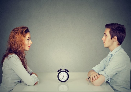 A man and woman sitting across from each other at a table with an alarm clock sitting on the table between them.